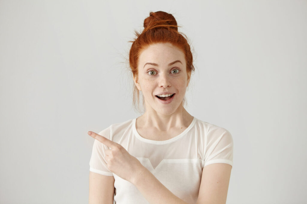 excited young redhead caucasian woman with hair knot pointing her index finger sideways raising eyebrows and keeping mouth wide opened showing something surprising