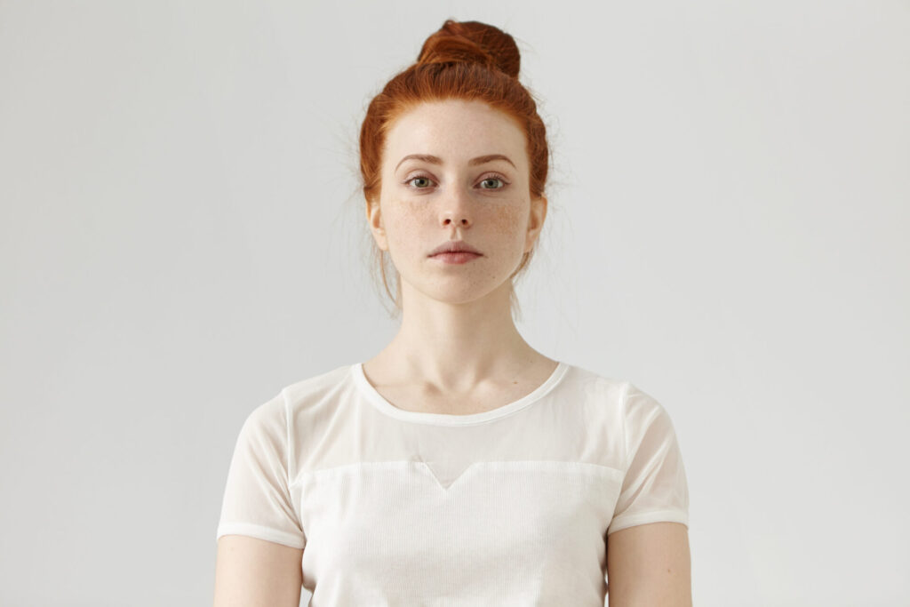 indoor portrait of attractive young european ginger woman with freckled face and hair bun dressed in white blouse her look and posture expressing self confidence